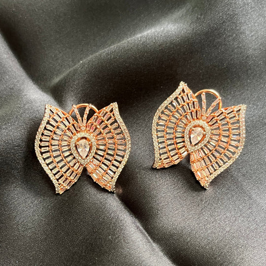 A pair of rose gold butterfly-shaped earrings with intricate filigree detailing and clear stones, displayed on a dark gray satin fabric background.